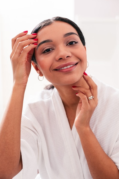 Heureux jeune femme dans un peignoir blanc souriant et touchant sa peau tout en posant pour une photo