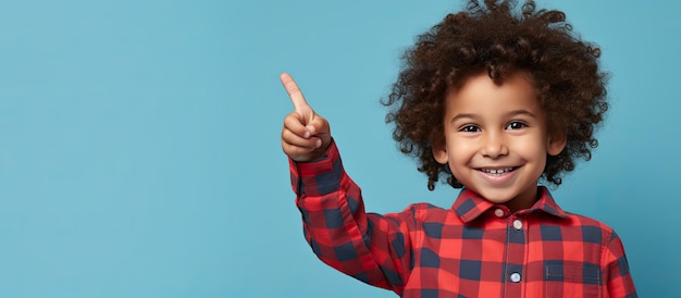 Heureux jeune garçon aux cheveux bouclés pointant vers le haut portant une chemise à carreaux rouge isolé sur fond bleu dans un portrait en studio