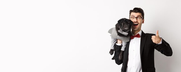 Heureux jeune hipster en costume et lunettes montrant le pouce vers le haut tenant un mignon chien noir sur l'épaule aime son