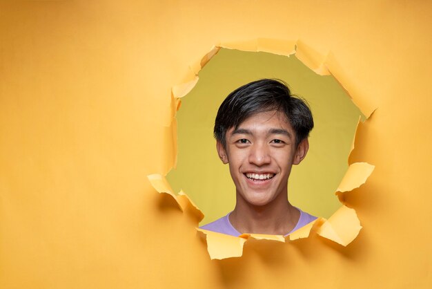 Heureux jeune homme adolescent asiatique souriant pose à travers un trou de papier jaune déchiré, portant un t-shirt violet avec un espace de copie ou du papier jaune vierge.