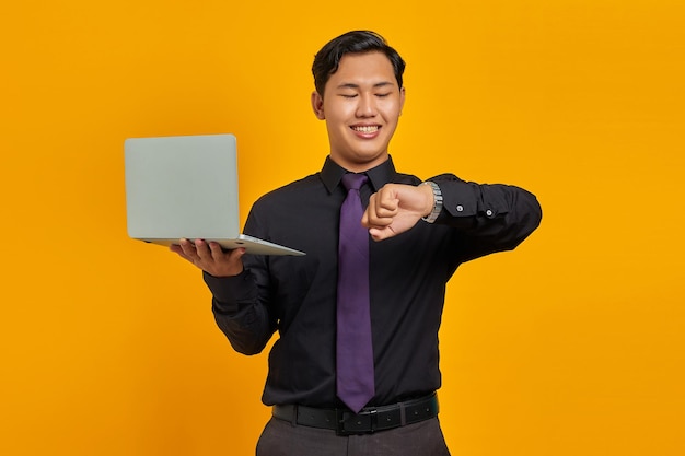 Heureux jeune homme d'affaires asiatique regardant une montre-bracelet et tenant un ordinateur portable isolé sur fond jaune