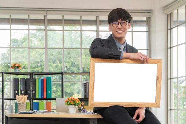 Heureux jeune homme d'affaires asiatique tenant un tableau blanc vide et assis sur la table au bureau.