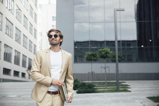 Heureux jeune homme d'affaires habillé en costume formel à la mode tenant un ordinateur portable à l'extérieur. Gars barbu en lunettes de soleil rondes noires debout près du centre d'affaires