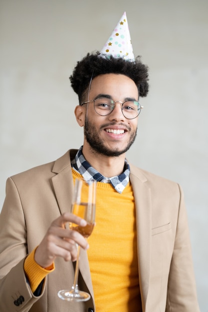 Heureux jeune homme d'affaires métis en tenue décontractée et casquette d'anniversaire grillant avec une flûte de champagne pétillant et vous regardant