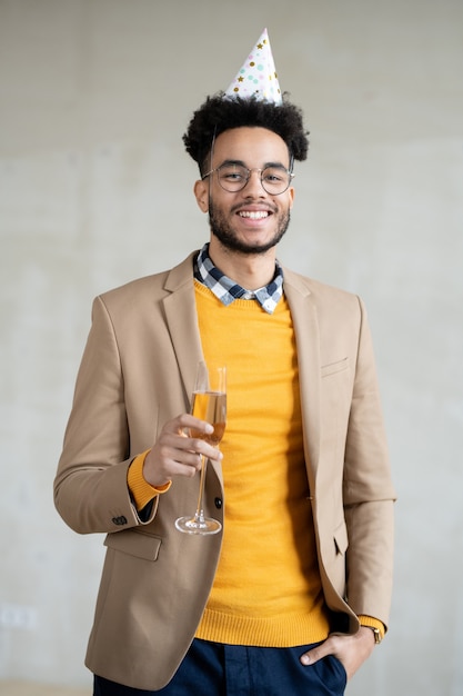 Heureux jeune homme d'affaires métis en tenue décontractée et casquette d'anniversaire tenant une flûte de champagne pétillant et vous regardant