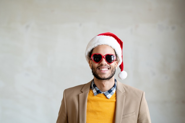 Heureux jeune homme d'affaires métis en vêtements décontractés intelligents, casquette de père Noël et lunettes de soleil en forme de cœur souriant tout en se tenant devant la caméra