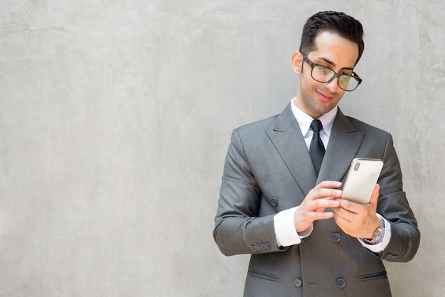 Heureux jeune homme d'affaires persan beau à l'aide de téléphone contre le mur de béton à l'extérieur