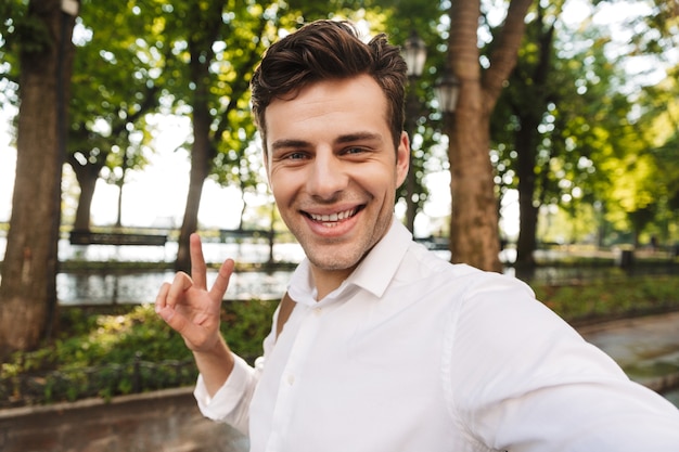 Heureux jeune homme d'affaires portant une chemise prenant un selfie en se tenant debout à l'extérieur dans le parc de la ville