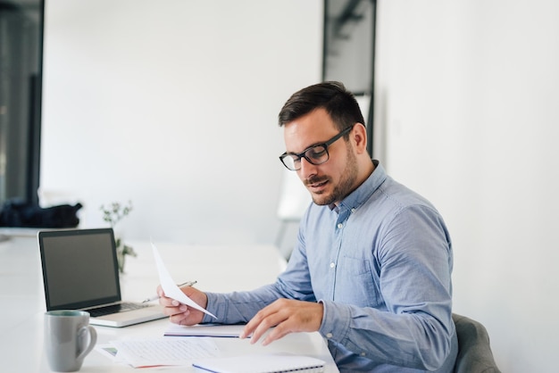 Heureux jeune homme d'affaires regardant des graphiques et des graphiques papiers à l'aide d'un ordinateur portable à son bureau