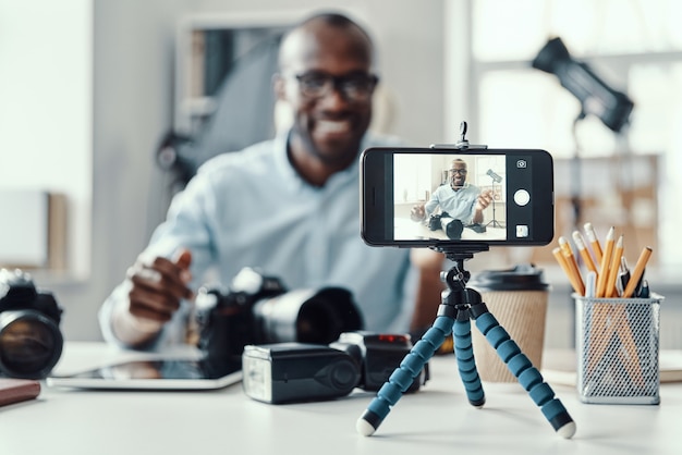 Photo heureux jeune homme africain en chemise montrant un appareil photo numérique et disant quelque chose tout en faisant une vidéo sur les réseaux sociaux