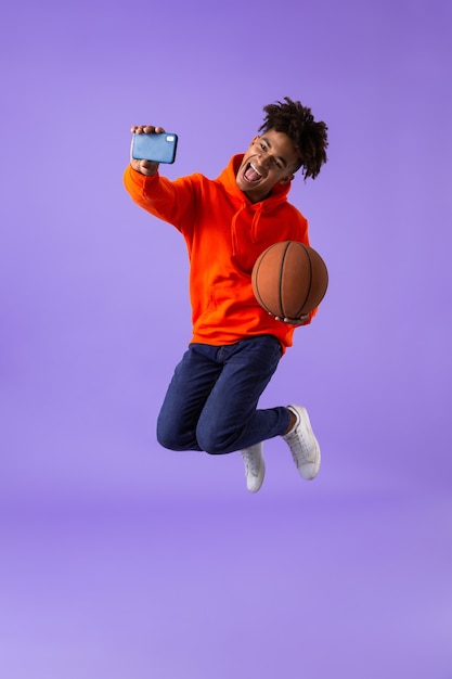 Heureux jeune homme africain posant isolé sur l'espace violet prendre un selfie par téléphone tenant le basket-ball.