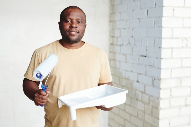 Heureux jeune homme africain avec rouleau à peinture et récipient en plastique blanc avec peinture debout contre deux murs dans son nouvel appartement ou maison