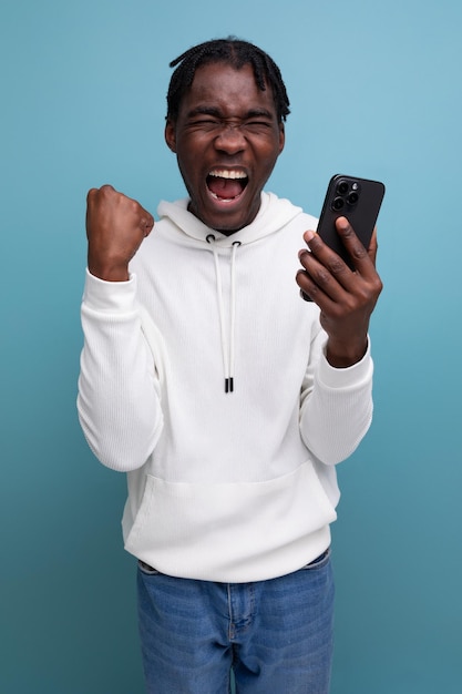 Heureux jeune homme africain en sweat-shirt blanc a remporté un concours en ligne à l'aide d'un téléphone intelligent