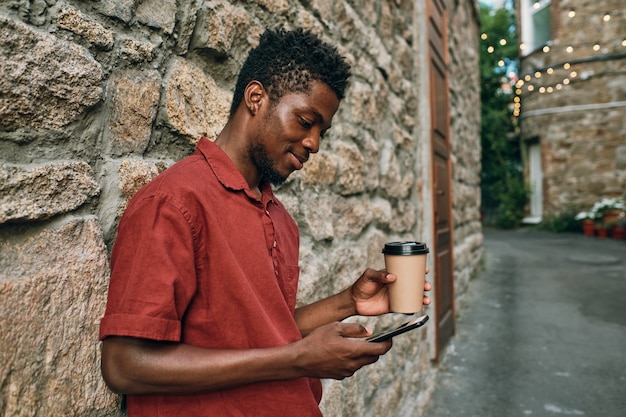 Heureux jeune homme africain avec des textos de boisson dans un smartphone