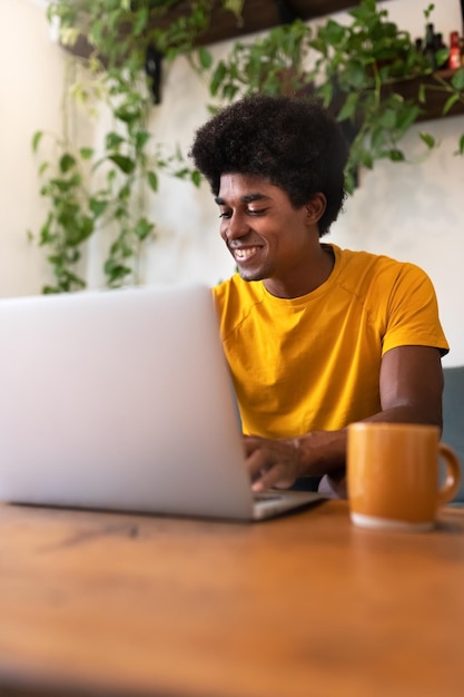 Heureux jeune homme afro-américain assis sur un canapé à l'aide d'un ordinateur portable le matin pour vérifier ses médias sociaux. Image verticale. Notion de technologie. À la maison.