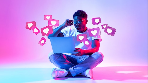 Heureux jeune homme afro-américain choqué avec la bouche ouverte enlève des lunettes a une conversation romantique avec des coeurs sur un ordinateur portable