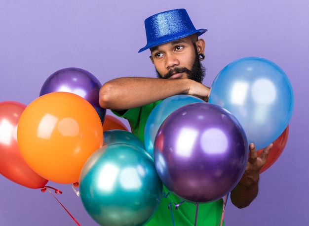 Heureux jeune homme afro-américain portant un chapeau de fête debout derrière des ballons isolés sur un mur bleu