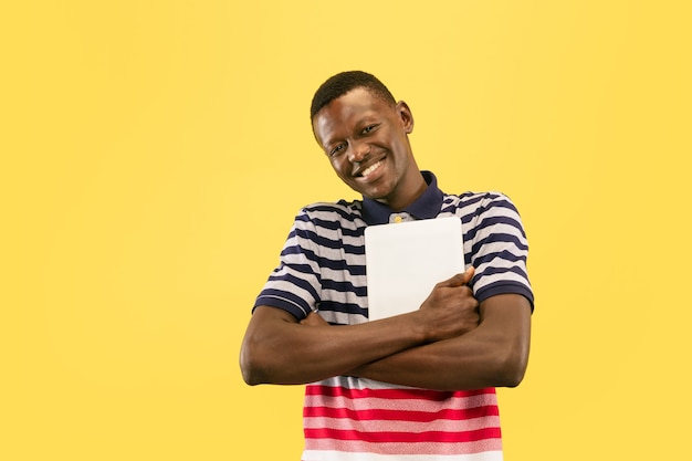 Heureux jeune homme afro-américain avec tablette isolé sur fond de studio jaune, expression faciale. Beau portrait d'homme mi-long. Concept d'émotions humaines, expression faciale.