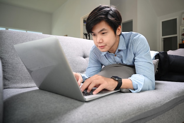 Heureux jeune homme allongé sur un canapé dans le salon et surfer sur Internet avec un ordinateur portable