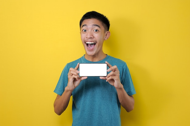 Heureux jeune homme asiatique excité montrant un écran de téléphone blanc à la caméra, isolé sur fond jaune