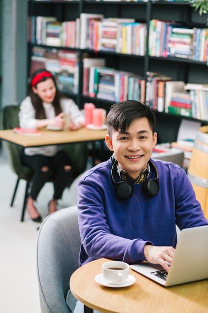 Heureux jeune homme asiatique mettant les mains sur le clavier de l'ordinateur portable et souriant alors qu'il était assis dans le cybercafé avec une tasse de café