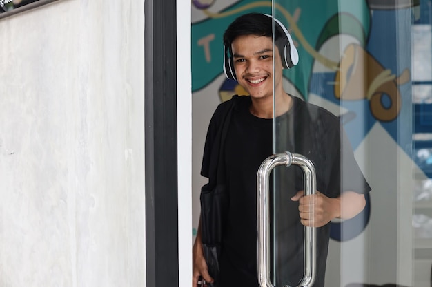 Heureux jeune homme asiatique portant des écouteurs sans fil souriant à la caméra tout en ouvrant la porte vitrée