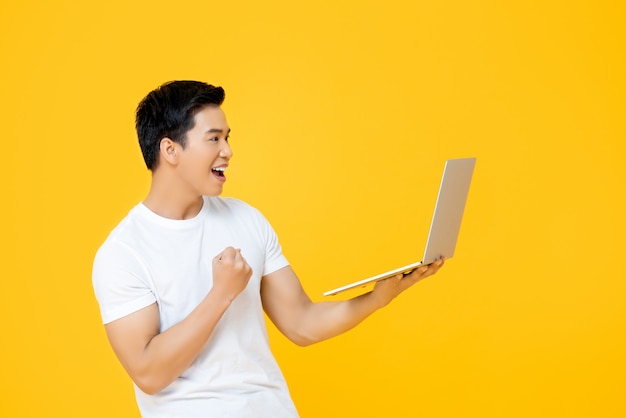 Heureux jeune homme asiatique regardant un ordinateur portable et levant son poing faisant oui geste isolé sur mur jaune