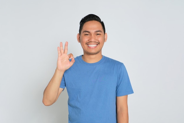 Heureux jeune homme asiatique en t-shirt bleu montrant un geste ok et regardant la caméra avec un visage souriant isolé sur fond blanc