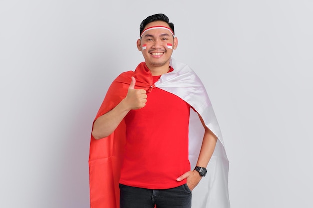 Heureux jeune homme asiatique tenant le drapeau indonésien tout en montrant les pouces vers le haut isolé sur fond blanc Concept de la fête de l'indépendance indonésienne