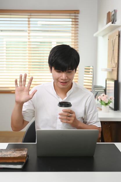 Heureux jeune homme asiatique tenant une tasse de café et saluant ses amis lors d'un appel vidéo sur un ordinateur portable.