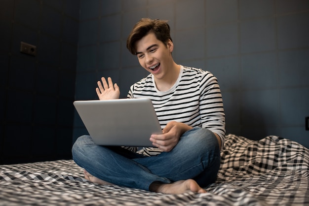 Heureux jeune homme assis sur un lit à l'aide d'un ordinateur portable