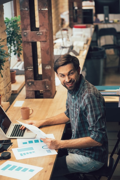Heureux jeune homme assis à la table du bureau avec des documents et se détournant de l'ordinateur portable avec un sourire