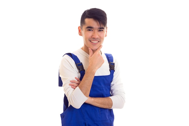 Heureux jeune homme aux cheveux noirs vêtu d'une chemise blanche et d'une combinaison bleue sur fond blanc en studio