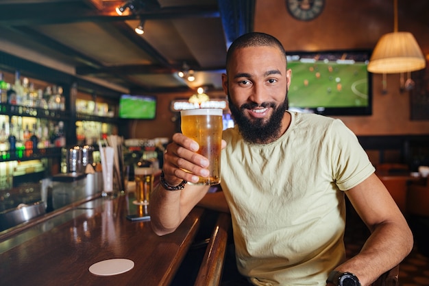 Heureux jeune homme barbu assis et buvant de la bière au bar