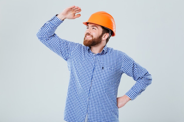 Heureux jeune homme barbu en casque debout et regardant au loin sur un mur blanc