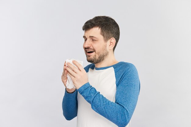 Heureux jeune homme barbu commence à éternuer dans son tissu blanc isolé sur mur gris