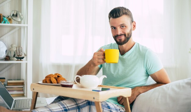 Heureux jeune homme barbu est assis sur son lit et prend son petit déjeuner avec thé ou café