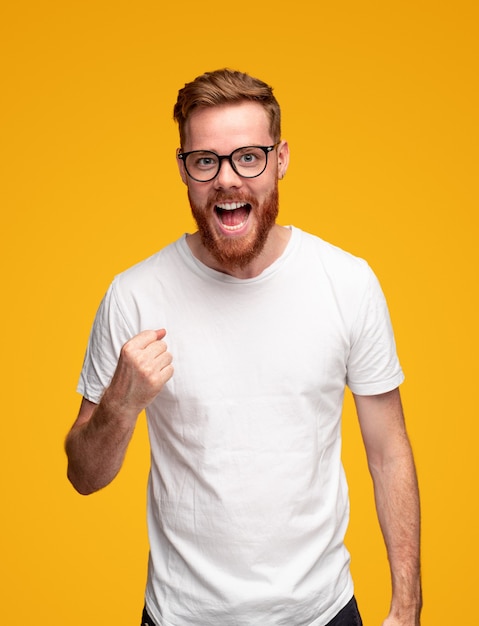 Heureux jeune homme barbu joyeux en t-shirt blanc et lunettes serrant le poing et criant tout en célébrant la victoire sur fond jaune