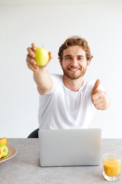 heureux jeune homme barbu à la table à la maison à l'aide d'un ordinateur portable tenant des fruits montrant le geste de pouce en l'air.