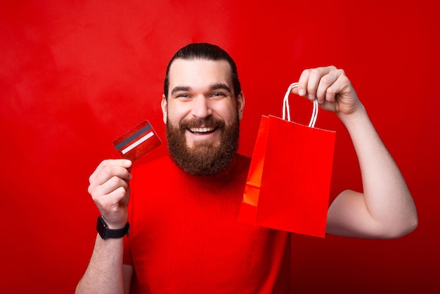 Heureux jeune homme barbu tenant sa carte de crédit et sac à provisions rouge, achat en ligne