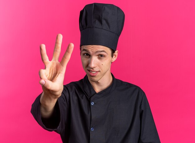 Heureux jeune homme blond cuisinier en uniforme de chef et casquette montrant trois avec la main