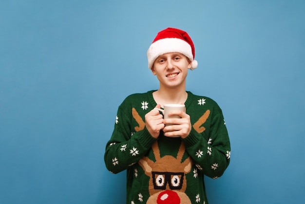 Heureux jeune homme en bonnet de Noel et pull de Noël avec des cerfs se dresse sur fond bleu