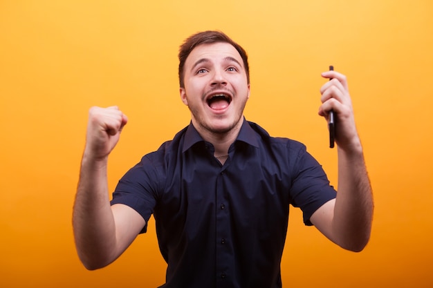 Heureux jeune homme en chemise bleue hondling téléphone portable et célébrer. Tourné sur fond jaune. Montrer de la satisfaction