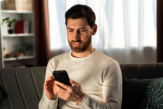 Heureux jeune homme communiquant par téléphone portable dans sa chambre pendant la soirée. Homme regardant l'écran avec un visage attentif