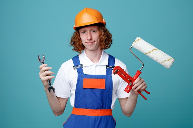 Heureux Jeune Homme Constructeur En Uniforme Tenant Des Outils De Construction Isolés Sur Fond Bleu