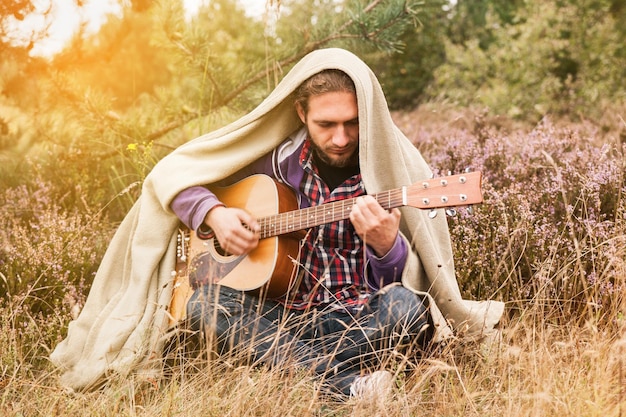 Heureux jeune homme couvert de plaid jouant de la guitare acoustique et chanter une chanson. Homme dans la forêt de pins avec pré de fleurs de bruyère. Gros plan, copiez l'espace.