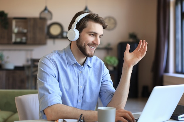 Heureux jeune homme dans des écouteurs travaillant sur un ordinateur portable depuis la maison pendant l'auto-isolement.