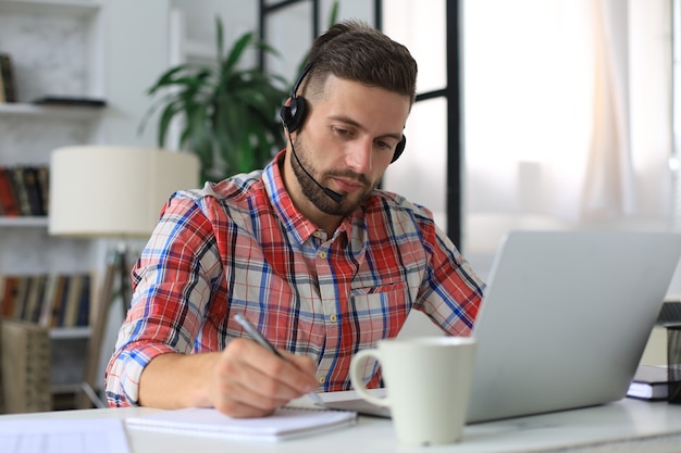 Heureux jeune homme dans des écouteurs travaillant sur un ordinateur portable depuis la maison pendant l'auto-isolement.