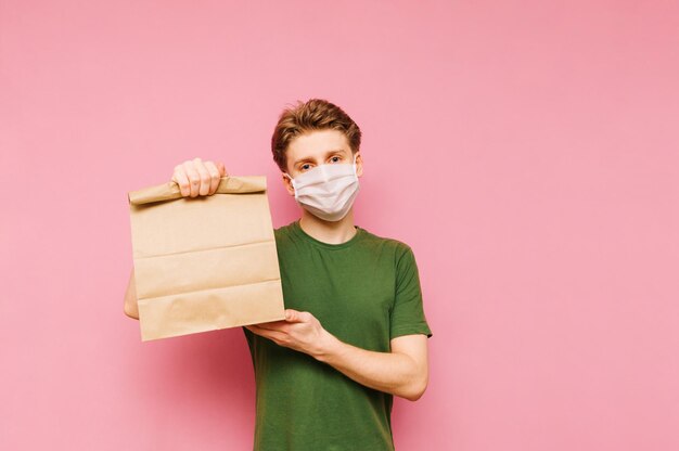 Heureux jeune homme dans un masque de protection se dresse sur un fond rose et tient dans sa main un sac en papier