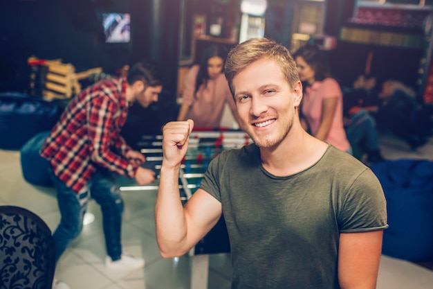 Photo heureux jeune homme debout dans la salle de jeux et pose devant la caméra. il sourit et tient la main dans le poing. ses amis jouent au baby-foot derrière.